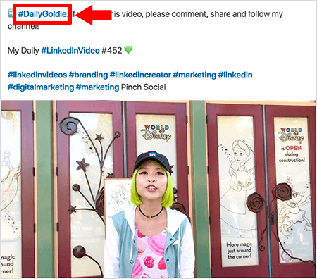 Bu, Goldie Chan'ın LinkedIn video gönderilerinin metninde hashtag'leri nasıl kullandığını gösteren bir ekran görüntüsü. Kırmızı belirtme çizgileri, metindeki #DailyGoldie etiketini işaret ediyor, bu da video gönderilerine özgü ve parça paylaşımlarına yardımcı oluyor. Gönderi ayrıca, #LinkedInVideo dahil olmak üzere insanların videosunu bulmasına yardımcı olan diğer ilgili hashtag'leri de içerir. Video görüntüsünde Goldie, World of Disney gösterisinde bazı kapıların önünde duruyor. Yeşil saçlı Asyalı bir kadın. Siyah bir LinkedIn şapkası, siyah bir gerdanlık kolye, pembe makaron baskılı bir gömlek ve mavi-beyaz bir ceket giyiyordu.